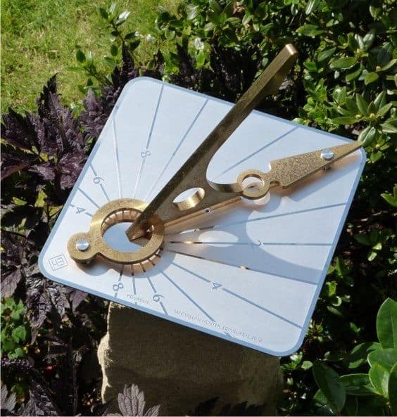 White sundial with brass-colored gnomon and hour lines against greenery backdrop.