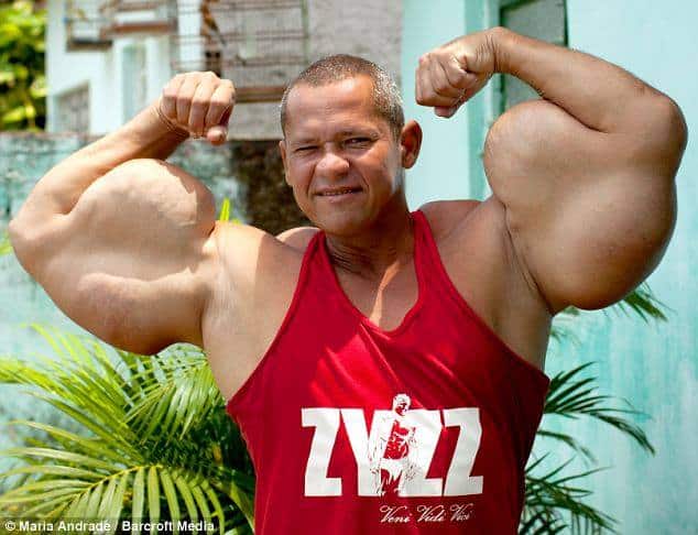 A man with short hair, wearing a red tank top, flexing his biceps in an outdoor setting.