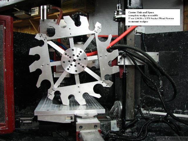 Metal machine device with central circle and six arm components on stand, with cables and machinery in the background.