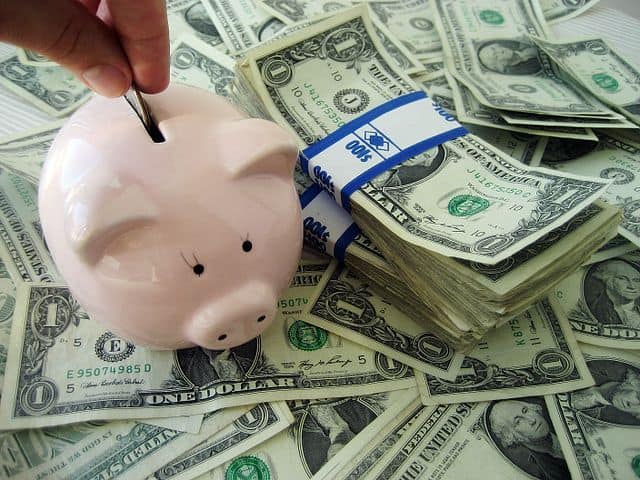A hand places money into a pink piggy bank with black eyes and a curved tail, surrounded by dollar bills.