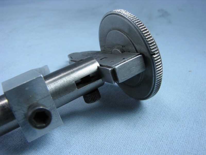 A dial indicator gage on a light blue background featuring a round base, circular knob, and extended stem with fine teeth and a small hole.