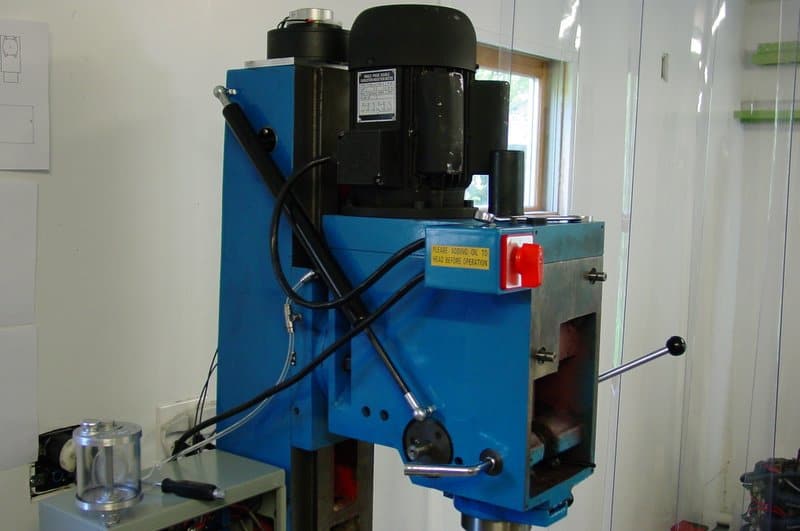 Blue metal drill press with black motor and attachments in a well-lit room.