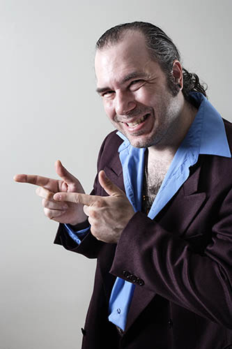 Man with slicked-back hair and sly grin, wearing dark suit jacket over light blue shirt.