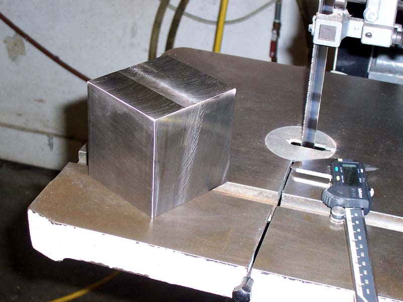 Precision instrument setup: stainless steel cube, digital caliper, metal rod with circular object on a white workshop background.