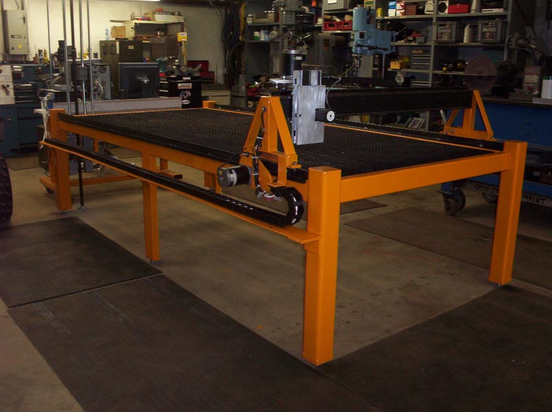 Workshop table with orange frame and black top, metal mesh surface and conveyor belt in a machine shop setting.
