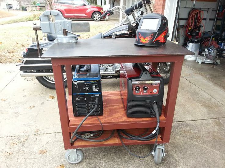 Portable welding cart with red frame and black top, featuring two welding machines and helmet.