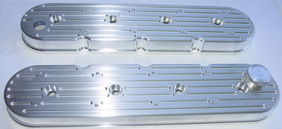 Two shiny silver valve covers with horizontal grooves, protruding bolts, and evenly spaced holes on either side, sitting on a white background.