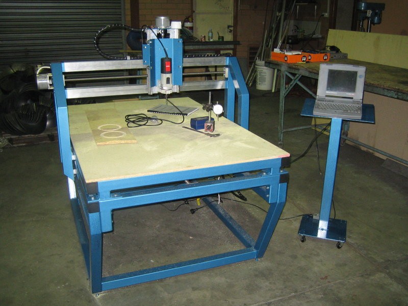 Blue and silver CNC router with laptop on top, large table, metal beams, and equipment in background.