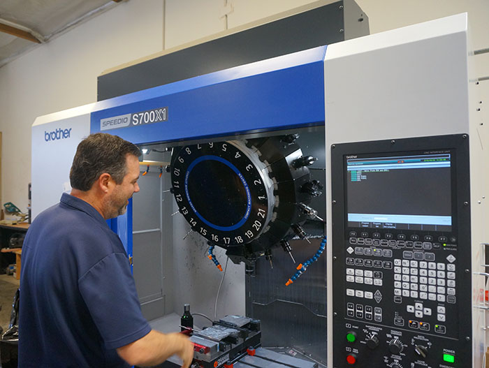 A man standing in front of a Brother machine with a circular scale labeled 1-21 attached to it, near a computer screen.