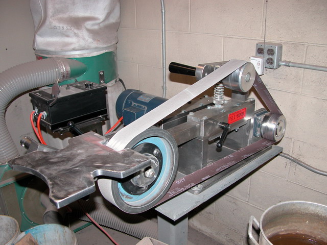 A metalworking belt grinder on an industrial workbench with green dust collector and gray hose.