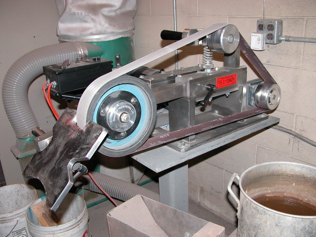 Industrial belt grinder with vacuum system on metal table against beige cinder block wall.