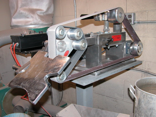A belt grinder with metal frame, silver components, red wires, and a gray hose sits against a cinderblock wall.