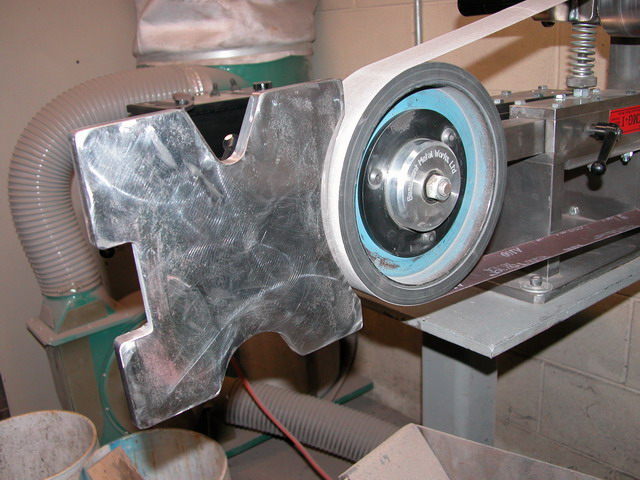 A metal belt sander attached to a machine with a black base plate and blue-edged serrated blade.