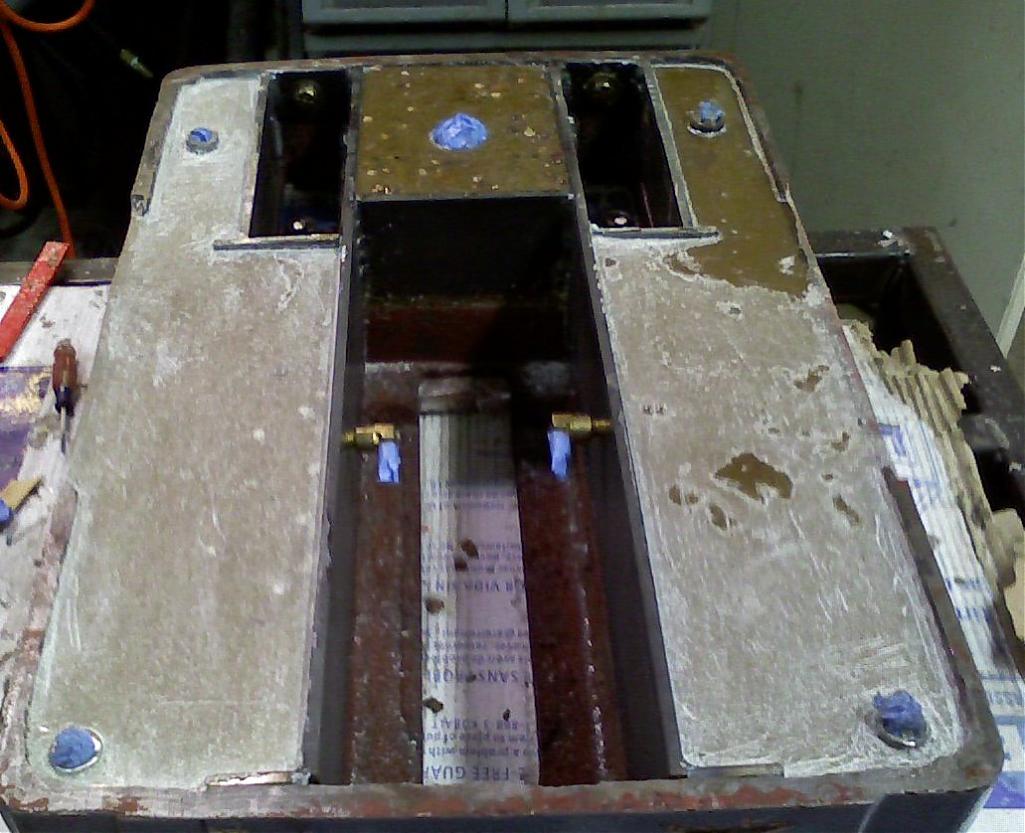 Metal base plate with cutout, grooves, and slots, featuring bolts on each corner and a white residue coating.