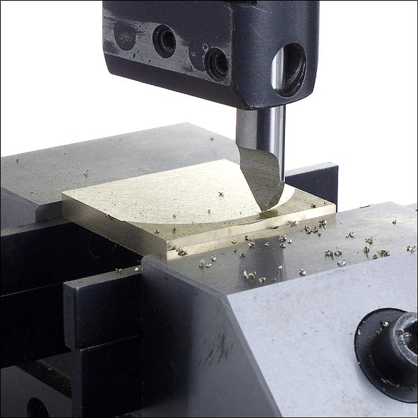 A sharp-edged tool attached to a machine holds a small piece of metal in place.