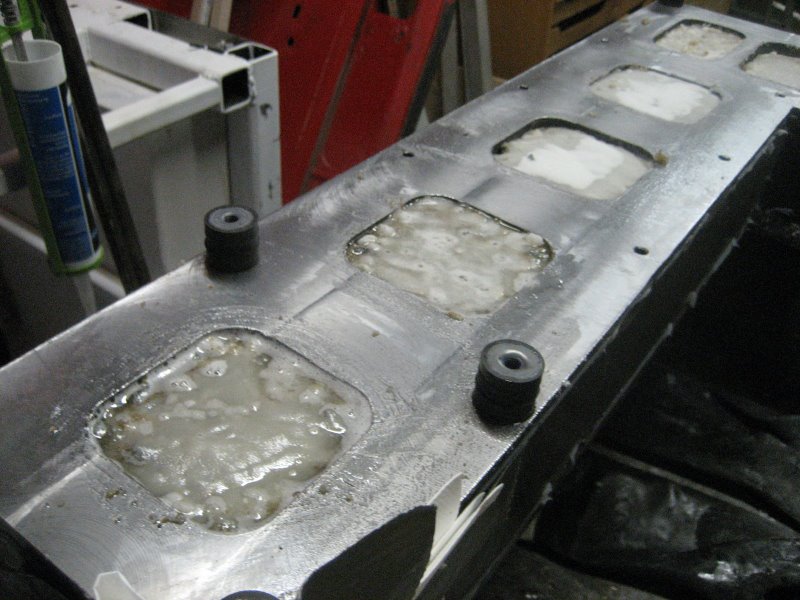 Metal component with applied substances and small square indentations containing dark or cylindrical features on a shiny silver surface surrounded by tools and materials.