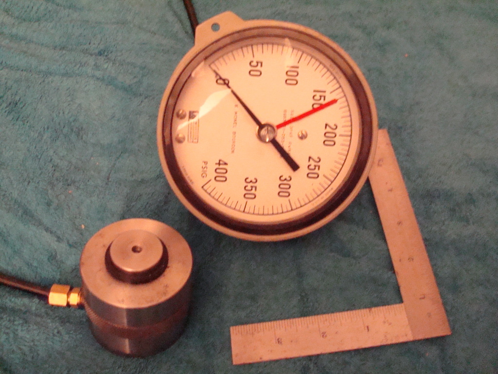 Large analog dial gauge on hydraulic press with black hose.
