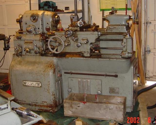 Rusted metal lathe with gear system and machinery controls in a cluttered workshop environment.