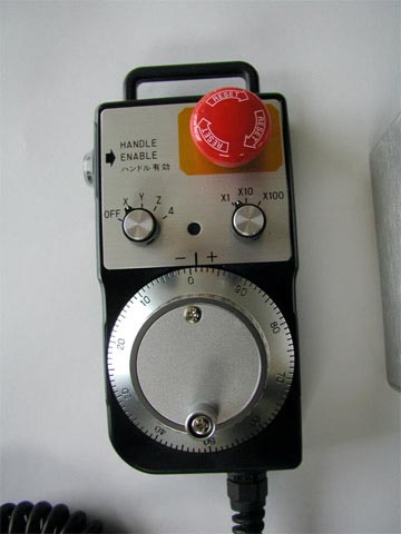 A control pendant with red button, white text, and multiple dials, made of metal or plastic, featuring a rectangular shape with rounded edges and a handle on top.