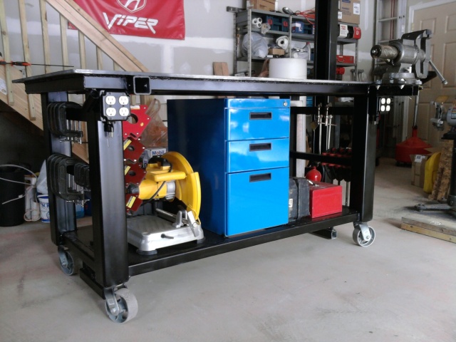 Large black rolling worktable with shelves, tool holders, press, drawers, and toolbox in a workshop/garage.