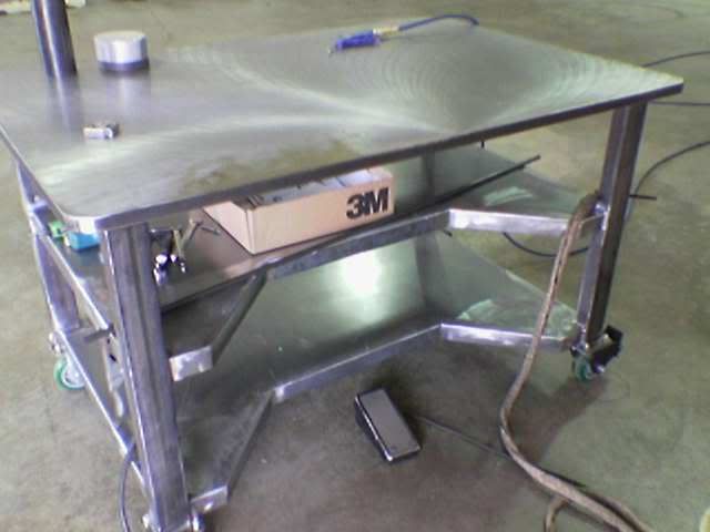 Stainless steel workbench with 1-inch thick plate and square legs featuring a shelf with a cardboard box and two small objects.