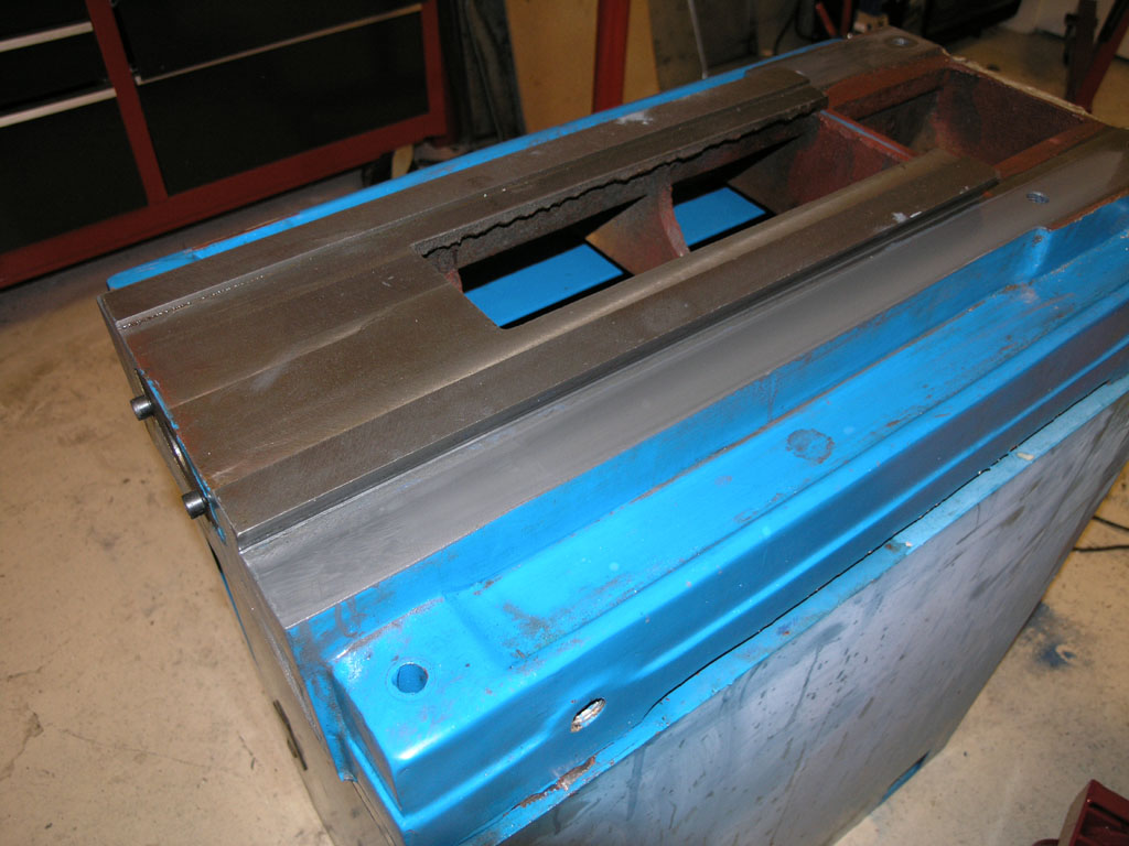 A large, rectangular metal object with blue paint and visible welds and bolts, set in a workshop or factory environment.