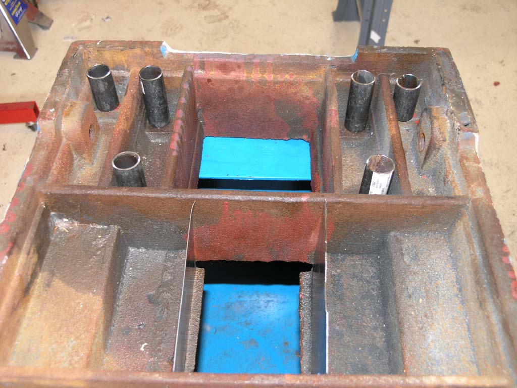 A metal frame with holes and cavities, brownish hue with red and blue accents, on a concrete floor.