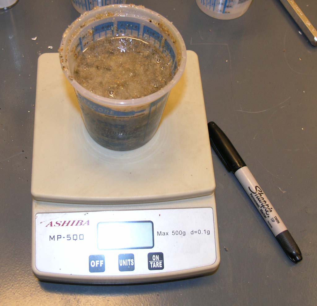 A laboratory scale with an unknown dark brown substance and a Sharpie marker in the background.