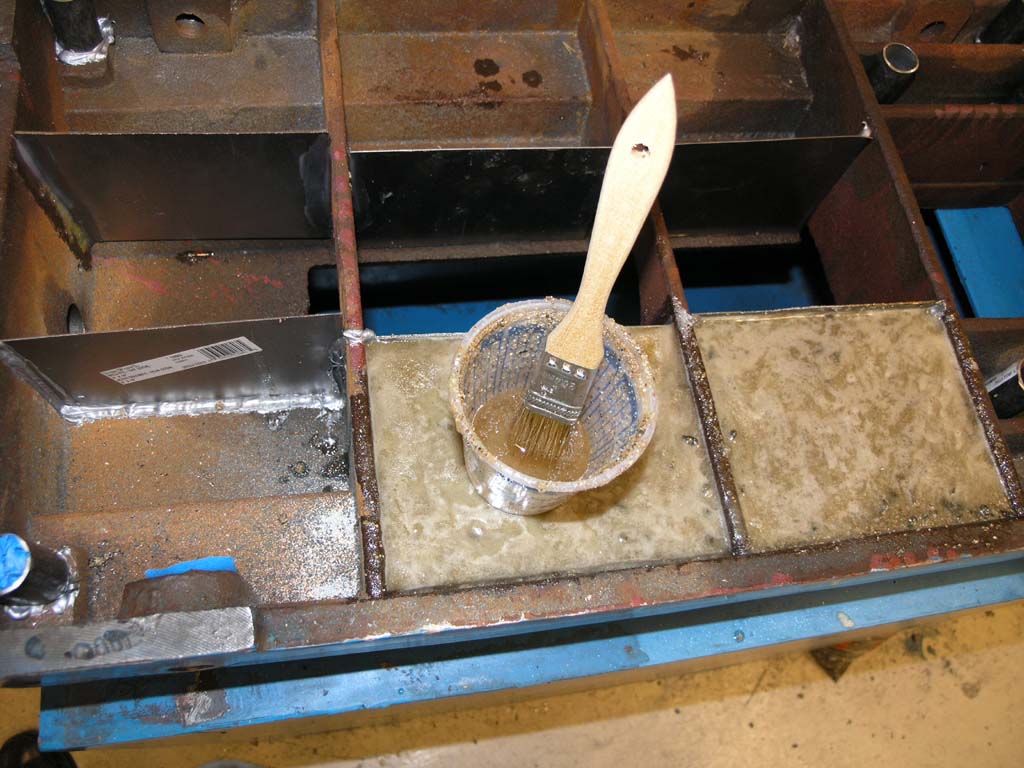 A rusty metal frame with attached metal pieces on a concrete floor, painted with brown liquid in a cup nearby.