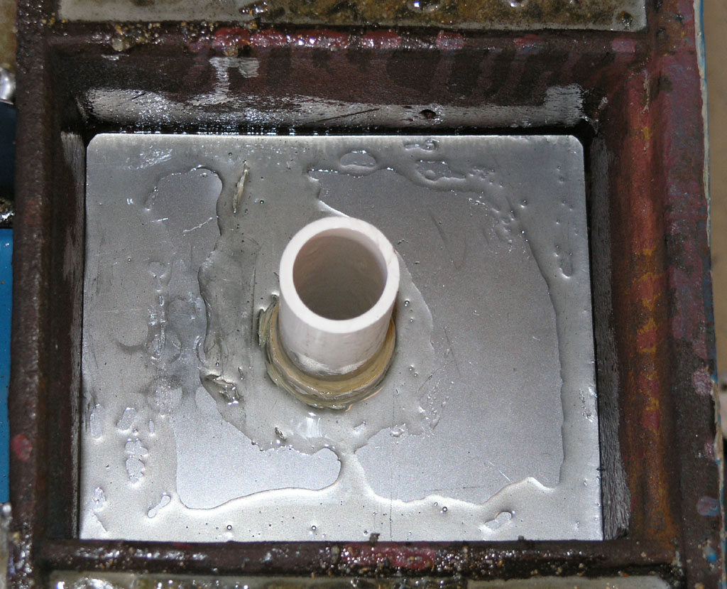 A square metal box with a white PVC pipe protruding from its center, surrounded by a dark brown rusted metal frame.