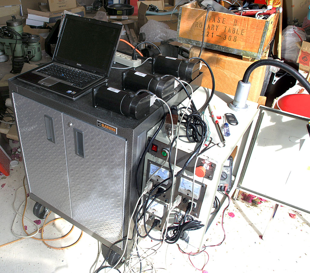 Cluttered workshop with tools and equipment scattered on the floor, featuring a large metal cabinet with silver finish and laptop on top.