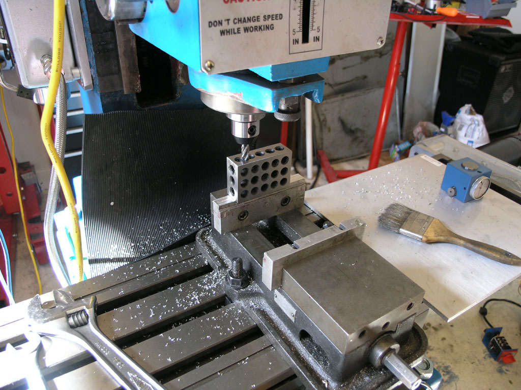 Blue and silver metalworking drill press or milling machine in workshop with table saw, control panel, large metal base, workpiece holder, and various tools and materials.