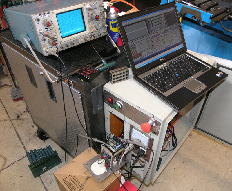 Cluttered workshop workbench with tools, laptop, oscilloscope, soldering iron.