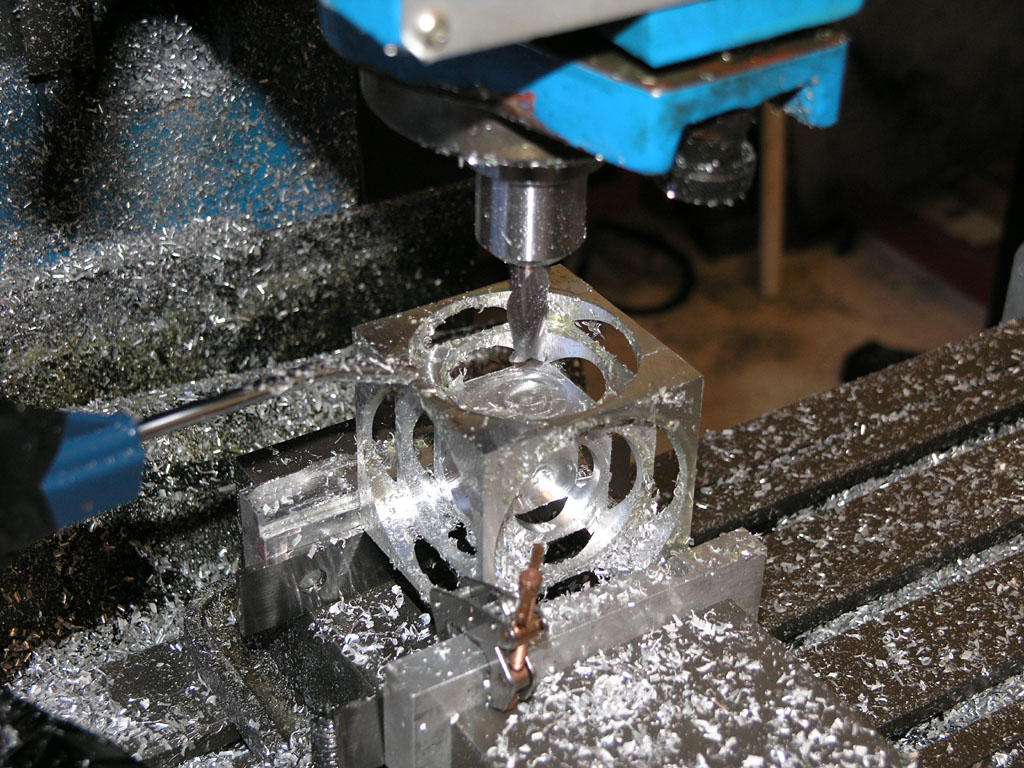 A CNC machined metal object with complex shape, hollow center, and curved cutouts, surrounded by shavings and debris.