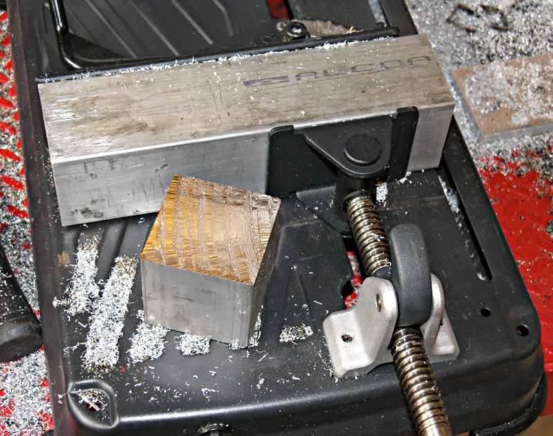 Metalworking vise holding a large metal part, surrounded by metal shavings on a red surface.