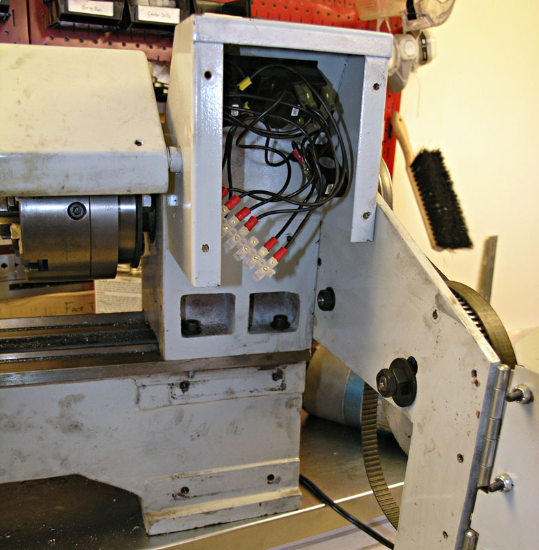 Old metal lathe with attached wires and tools, positioned in front of a wall with a pegboard behind.