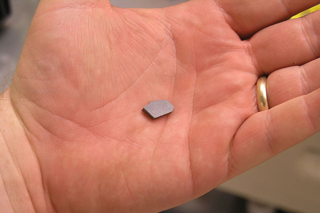 Man's hand with gold wedding band holding small gray metallic or plastic object.