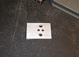 Rectangular metal plate with three circular holes on dark gray surface.