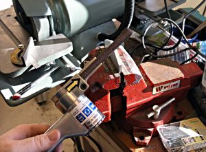 Hand holding tool in front of red metal vise on workbench with yellow arrow and black cord.