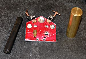 Red metal workbench clamp with black handle and unmarked brass object against dark gray background.