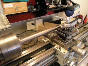 Metal lathe with cylindrical workpiece being turned, surrounded by various tools and parts in a metalworking shop setting.