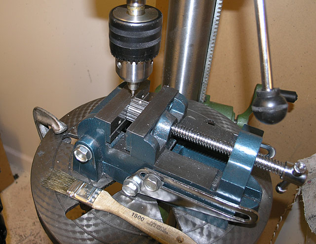 A blue metal bench vise with adjustable jaws securely holding a drill bit and an object.