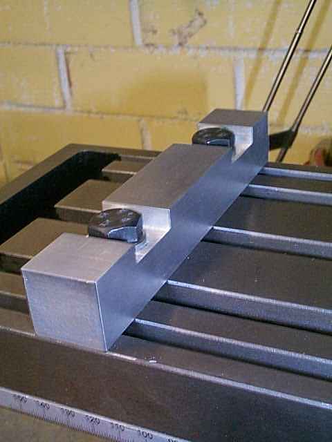 Dark gray metal vise with perpendicular edge clamp and black lever, set against a yellow brick wall.