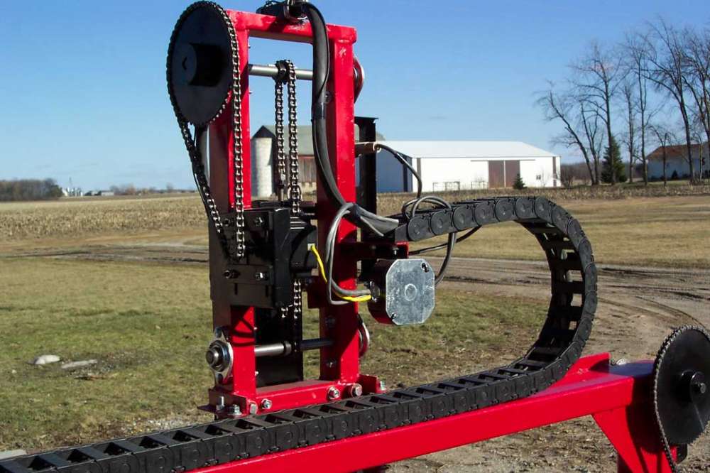 Red metal machine frame with wheels and track in a field background with a white barn.