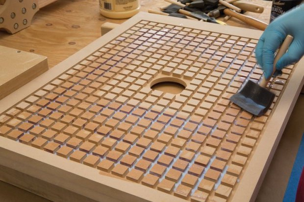 Person in blue gloves applying protective coating to wooden object with small squares and central hole.