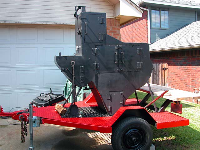A large black smoker shaped like a pig sits atop a red trailer in front of a brick house.