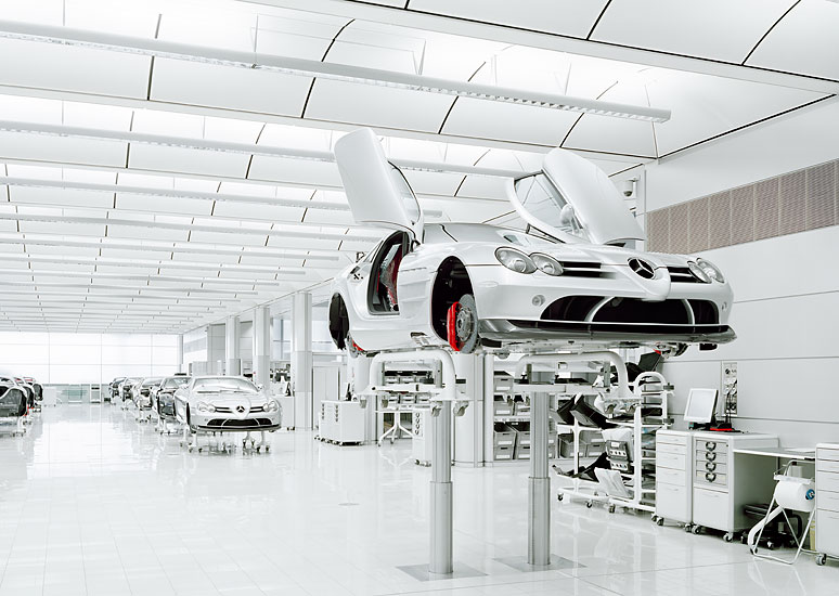 A workshop or factory setting with white walls and floors, featuring cars on stands or lifts and various machinery.