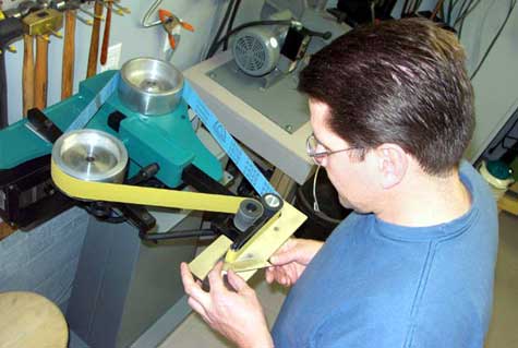 A person in a blue shirt adjusts a tool on a horizontal grinder in a cluttered workshop.