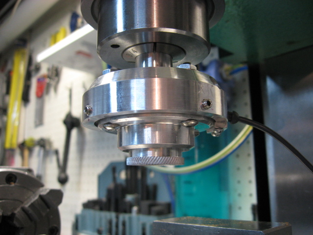 A silver drill press spindle on a blue stand against a white pegboard background with tools and accessories.
