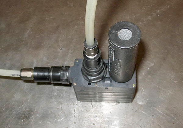 Small, cylindrical device on aluminum block with hole and white tube connection.